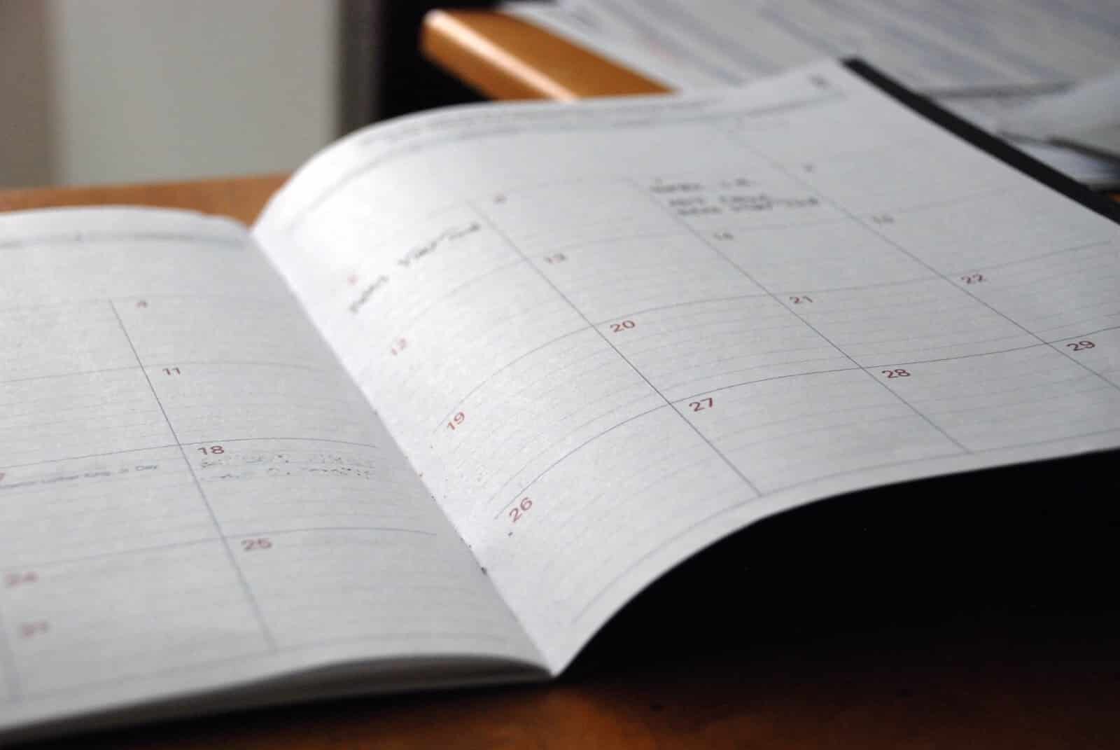 An open planner displaying a monthly calendar layout is resting on a wooden surface. The dates are visible, but no specific appointments or notes are legible. The planner appears to be partially filled with handwritten entries. In the background, blurred documents are visible.