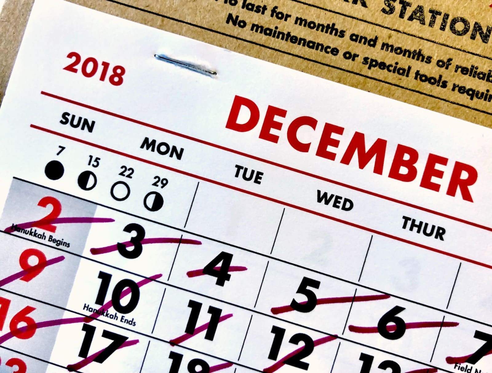 A close-up of a 2018 wall calendar showing the month of December. The 7th is marked as "Hanukkah Begins", while the 10th is labeled "Hanukkah Ends". Several dates including the 3rd to the 7th have been crossed out with a red X.