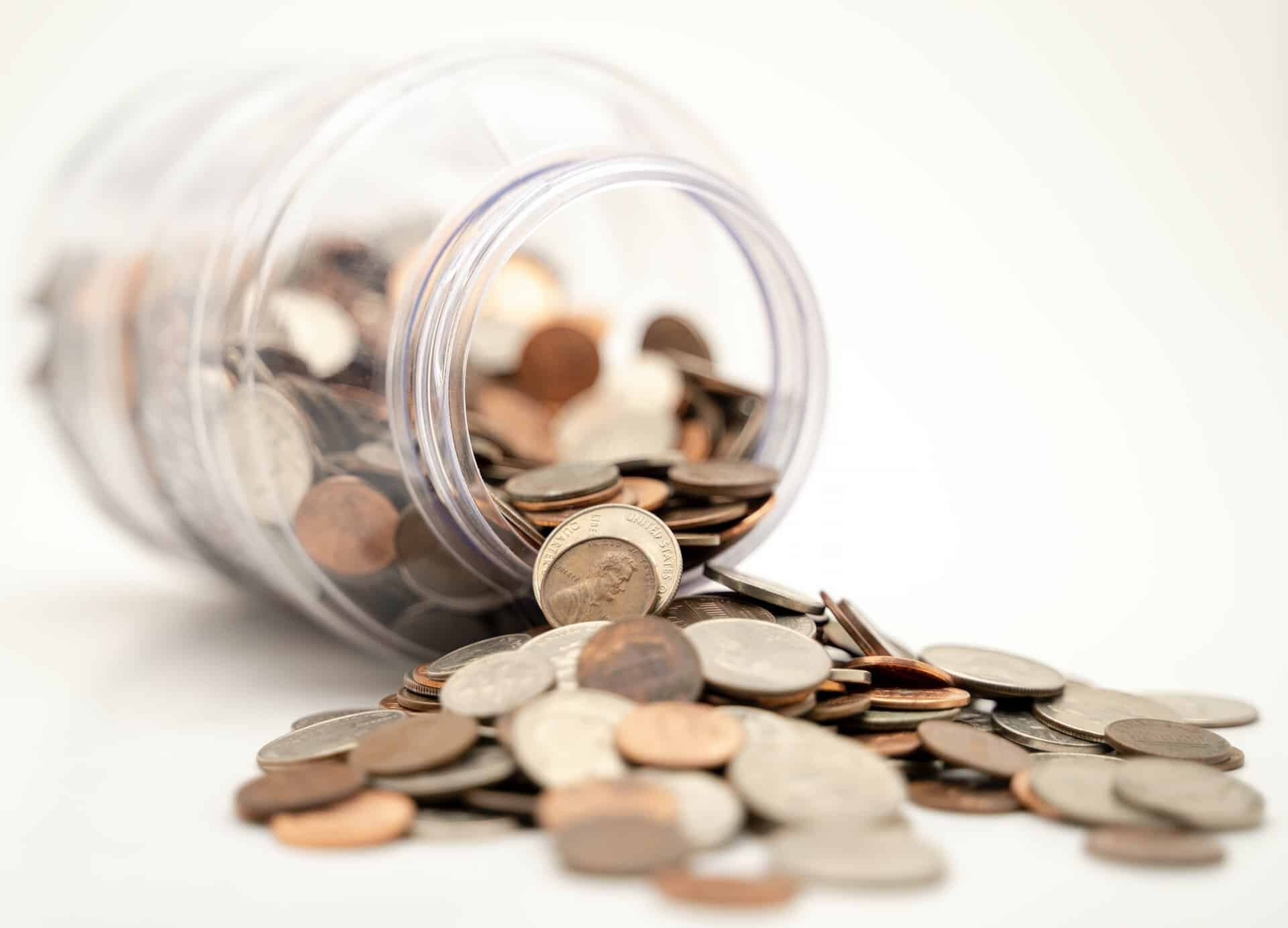 A clear plastic jar tipped over, spilling a variety of coins, including nickels, dimes, and pennies onto a white surface. The coins correlate with the 2021 HTF Income Limits, now scattered around the overturned jar.