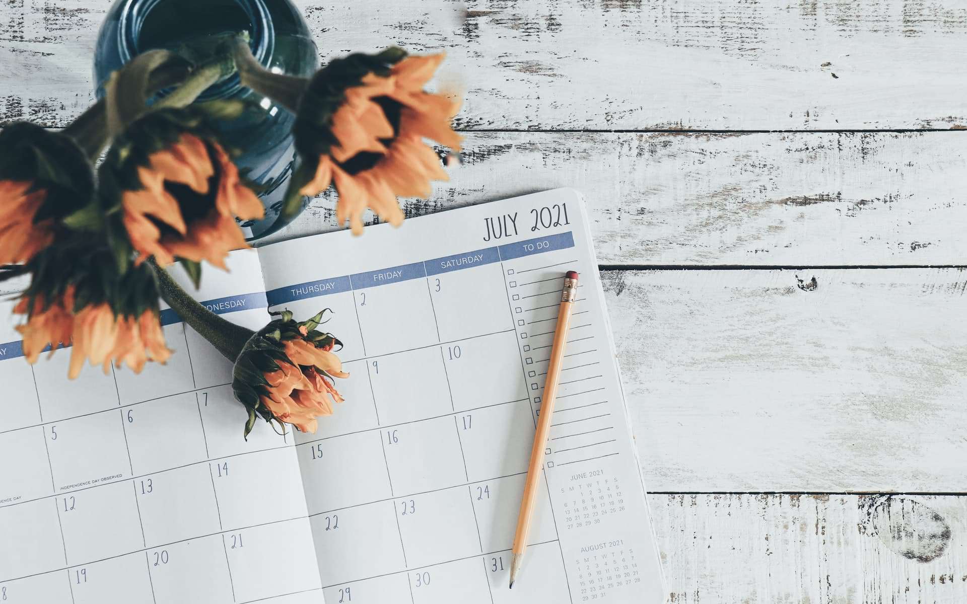 A calendar is opened to July 2021 on a white, distressed wooden surface. An orange pencil is placed diagonally on the calendar, and a blue vase with orange flowers is positioned on the left side of the image.