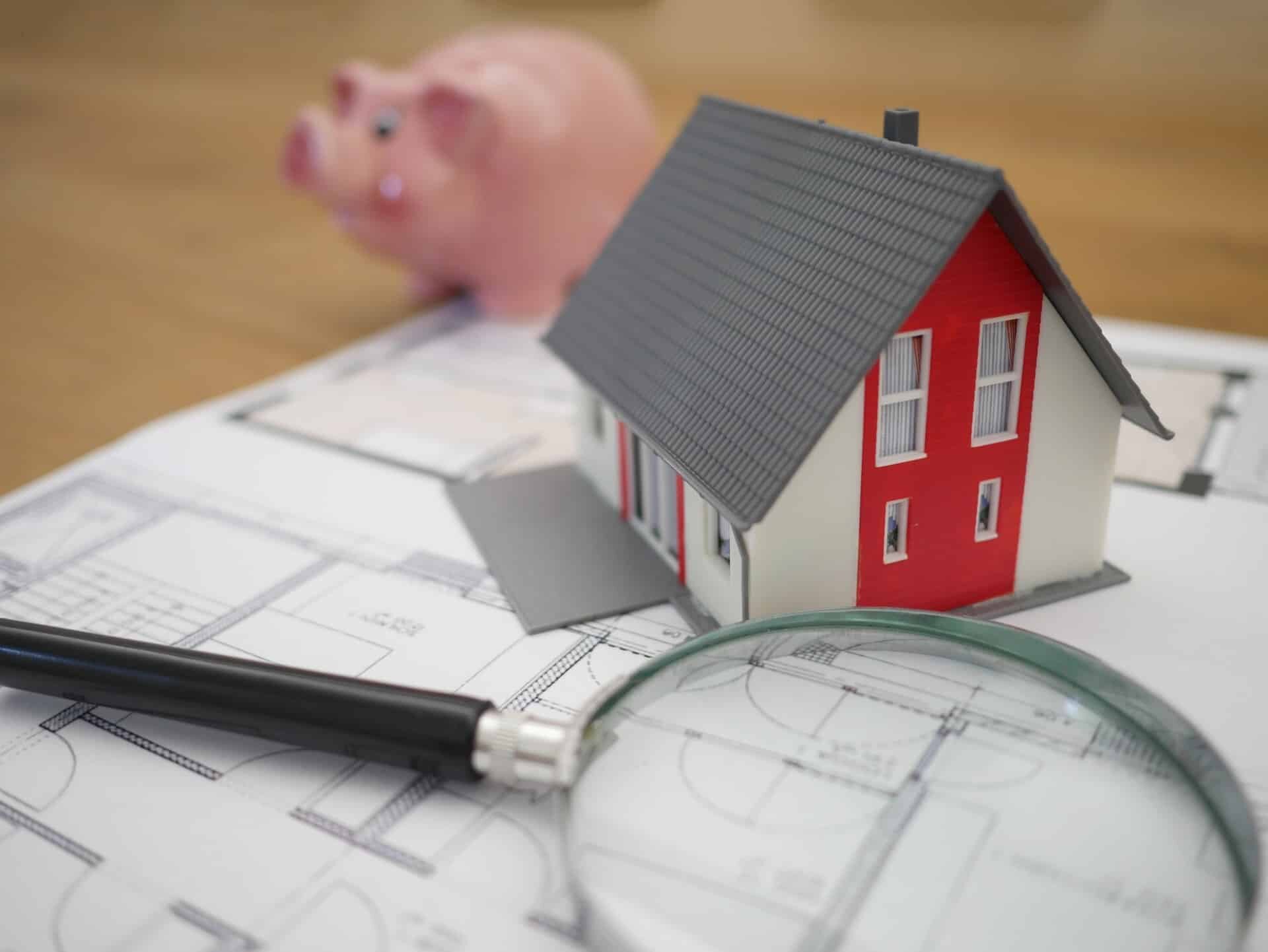 A miniature house model with red and white walls sits on architectural blueprints labeled "2022 HOME Limits." Nearby, a black pen and a magnifying glass rest, while a pink piggy bank is in the background, slightly out of focus.