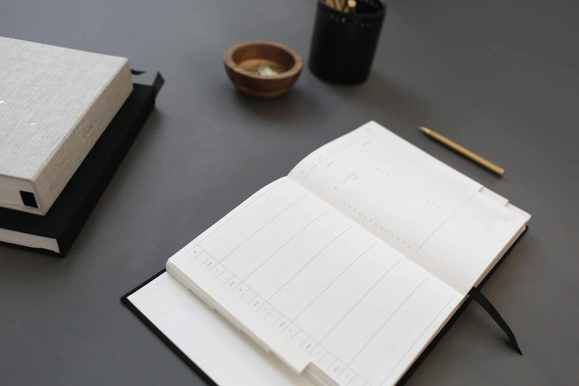 An open planner lies on a dark desk, displaying blank pages. To the left are two closed books, one with a gray cover marked "2022 MA EOY Submissions." A bowl and a container holding pencils are also on the desk, with a gold pen lying to the right of the planner.