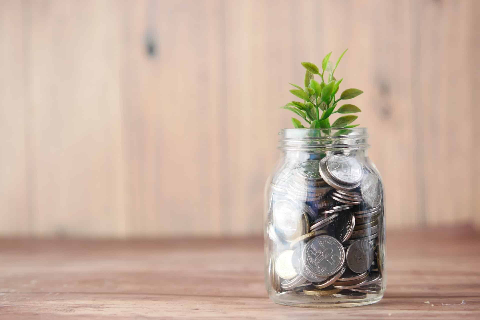 A small glass jar filled with silver coins has a young green plant growing out of it. The jar is placed on a wooden surface with a warm, blurred wooden background, symbolizing savings, investment, and growth.
