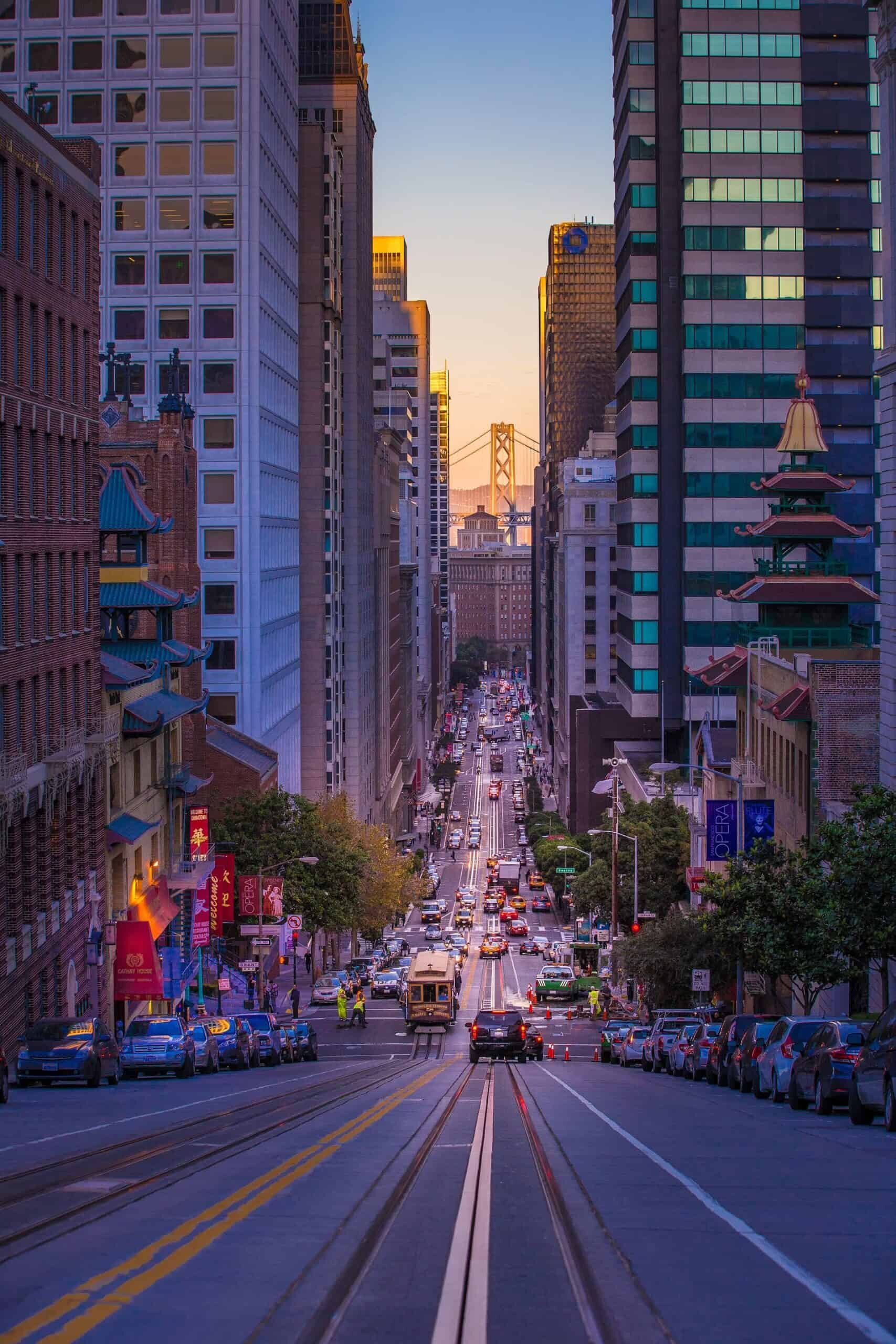 A bustling urban street scene at dusk features a steep hill lined with tall buildings on both sides. Cars and a cable car travel down the road, leading the eye to a distant bridge illuminated by the setting sun. Street banners and trees decorate the scene, subtly hinting at 2021 CA HUD Tenant Data Collection themes.