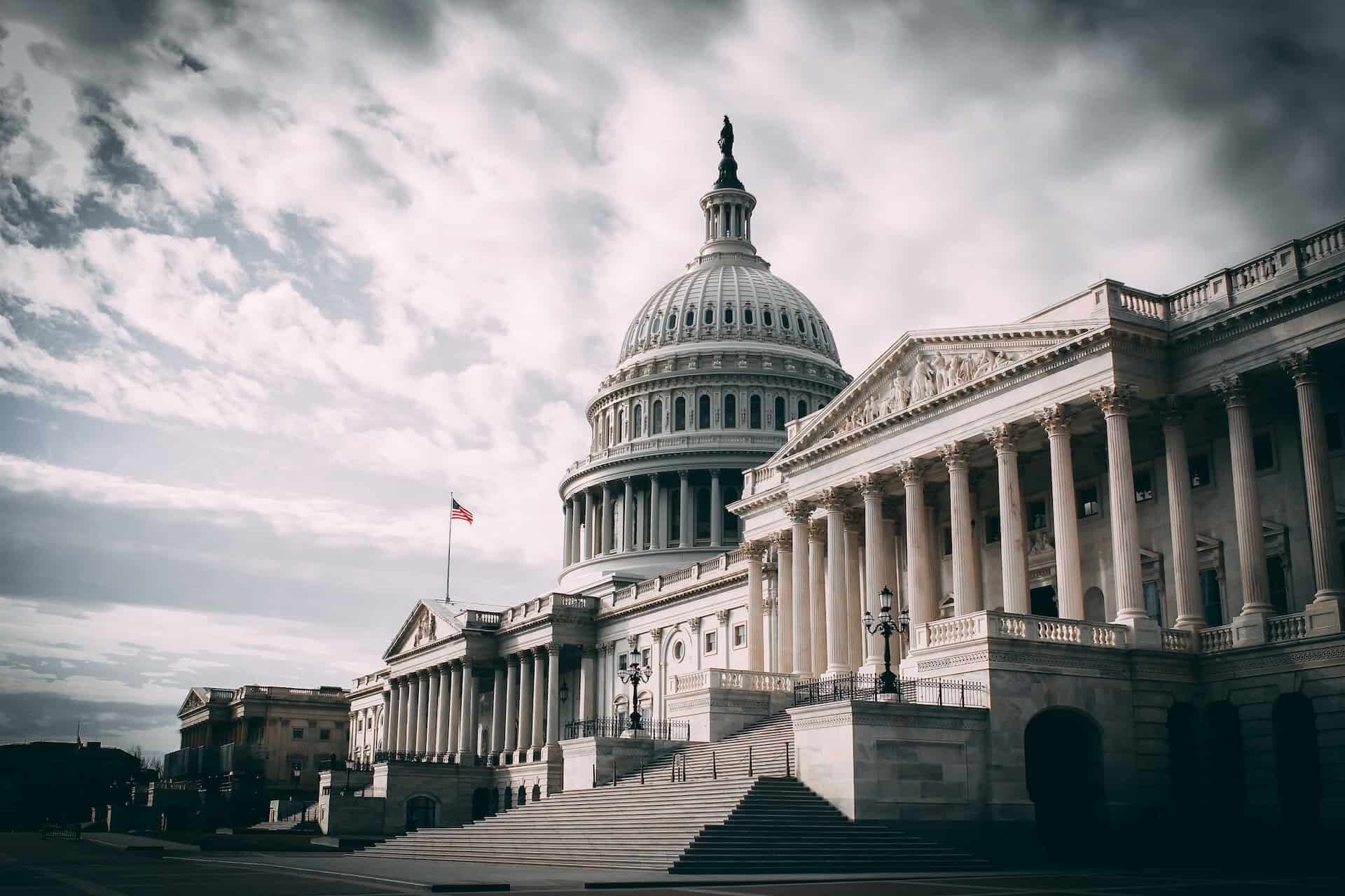 A large neoclassical building with a prominent white dome topped with a statue stands majestically. The building features many columns and a wide staircase leading to the entrance, where discussions on the 2023 Income Limits take place. An American flag is visible near the dome against a cloudy sky.