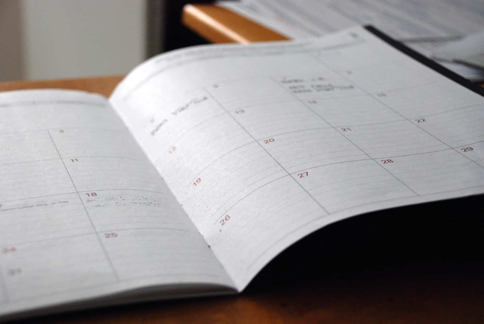 An open planner or calendar lies on a wooden desk, showing a monthly view with numbered dates. The planner has some handwritten notes on certain dates, and a stack of papers can be seen in the background, slightly out of focus.