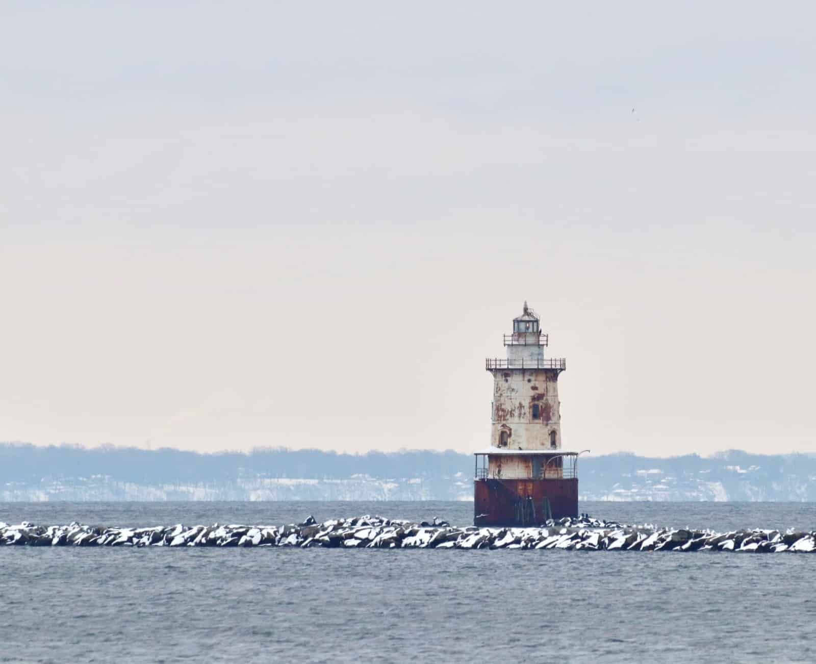 A small lighthouse standing on a frozen lake with snow-covered ground and distant hills in the background