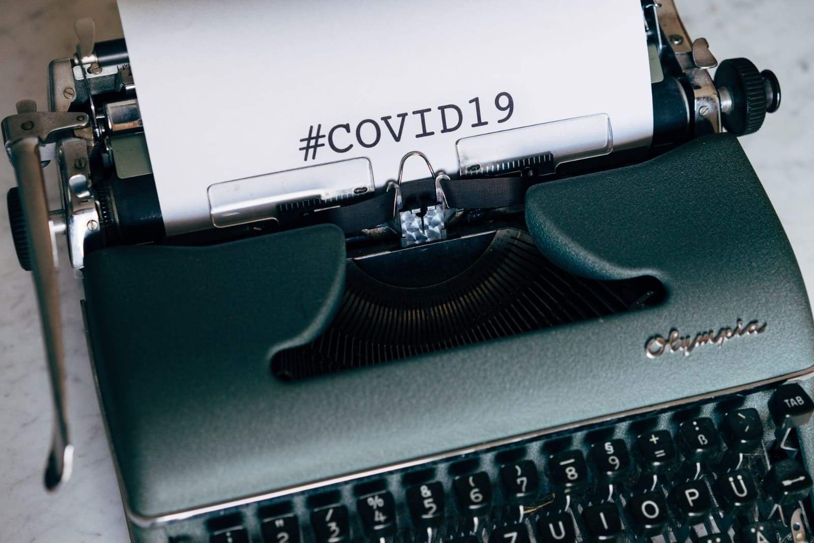 A green typewriter with a sheet of paper inserted displaying the text "#COVID19." The typewriter is vintage in style, with the brand name "Olympia" visible on the front. The paper appears to have been freshly typed with the hashtag phrase.