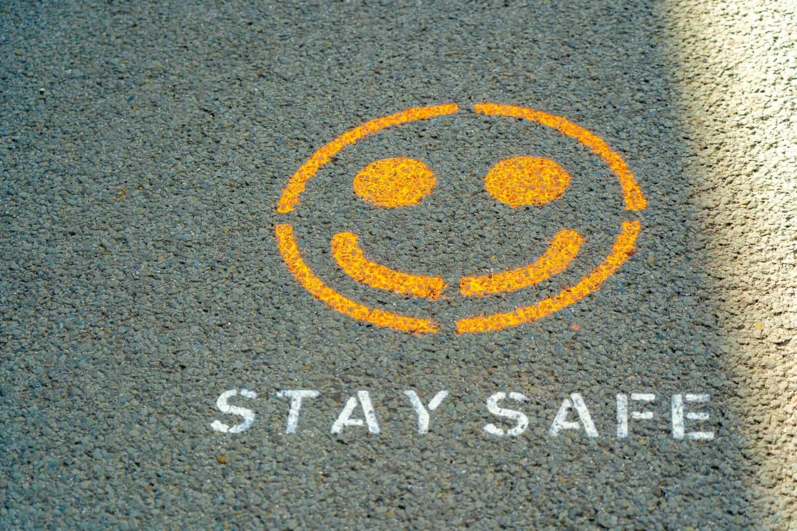 A yellow smiley face is painted on a gray pavement, with the words "STAY SAFE" written in white underneath. Sunlight partially illuminates the scene.