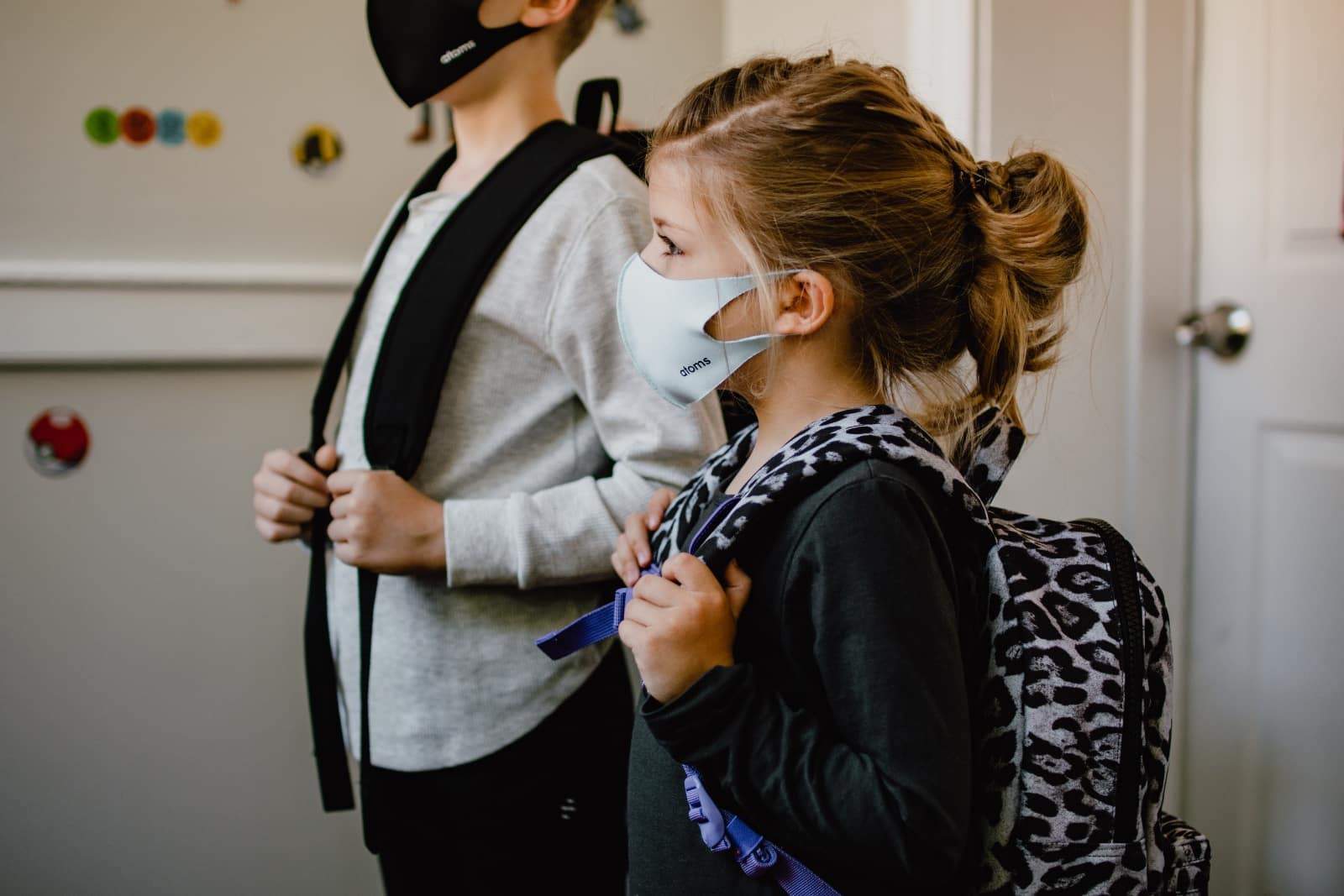 Two children wearing face masks, standing indoors with backpacks. The child in the foreground has a ponytail and wears a patterned backpack, while the child in the background has short hair and a plain backpack. Both are dressed in casual clothing.