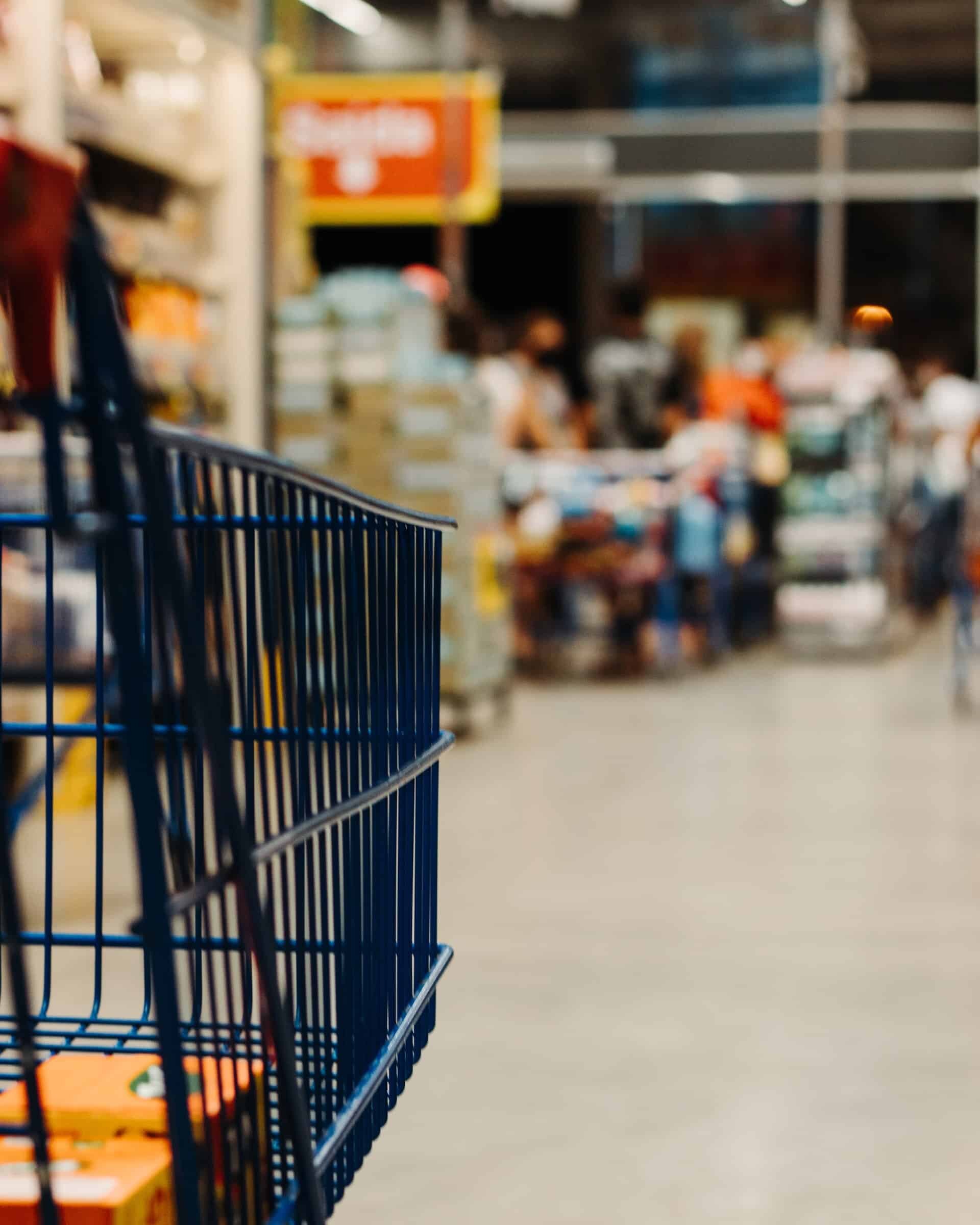 Close-up of an empty blue shopping cart in a grocery store aisle. In the background, shelves are stocked with products, and people are visible further down the aisle, slightly blurred. The scene is brightly lit, capturing a busy Walmart environment amid the recent minimum wage increase buzz.