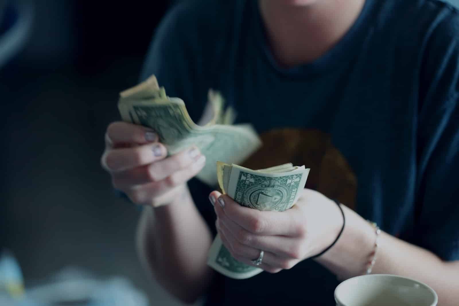 A person is counting a stack of U.S. dollar bills, holding some in each hand. The person is wearing a blue shirt and has a ring on their left hand. The image is taken close-up, focusing on the hands and the money. A mug is partially visible in the bottom right corner.