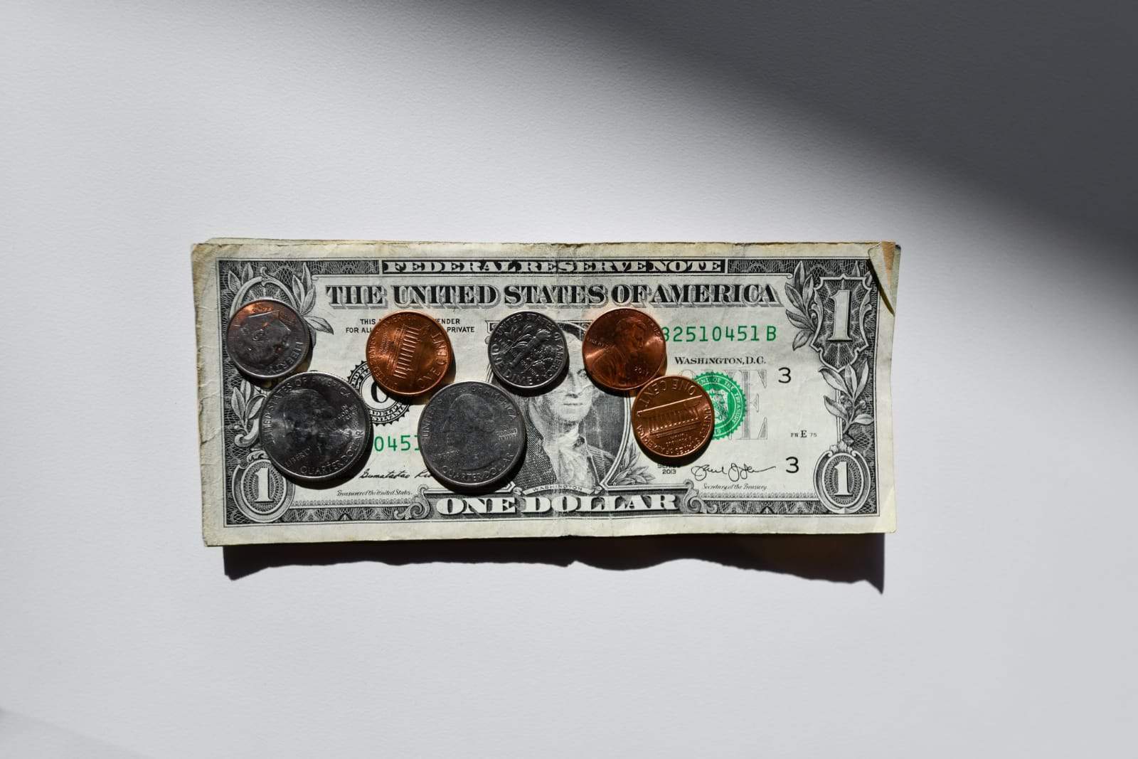 A crumpled one-dollar bill is laid flat on a white surface. Several coins, including pennies, nickels, dimes, and quarters, are scattered on top of the bill. The scene is lit by sunlight, creating shadows around the objects.
