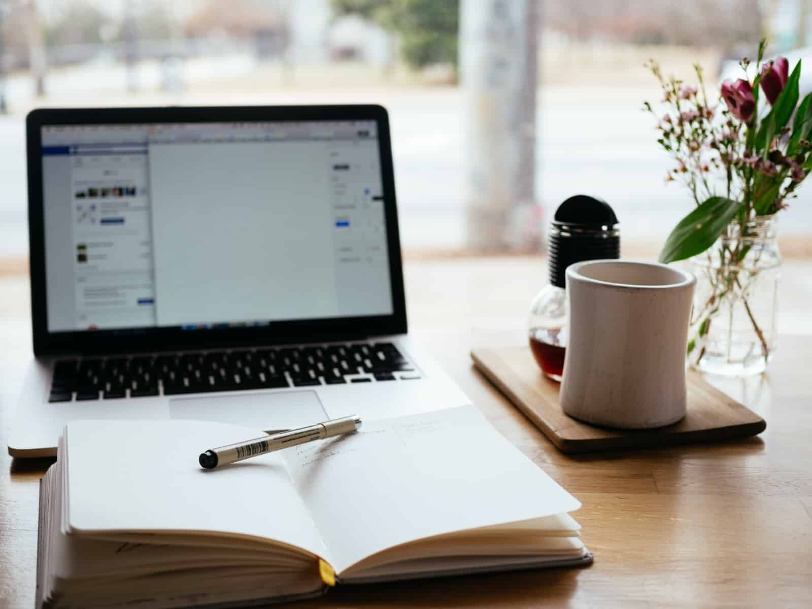 A workspace features an open laptop, an open notebook with a pen resting on it, a coffee cup on a wooden tray, and a small vase with flowers. The background shows a blurred outdoor scene through a window.