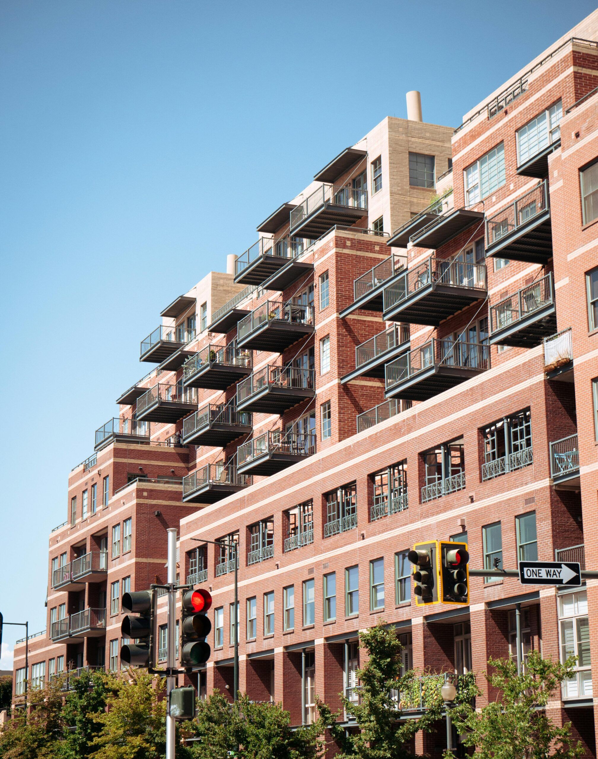 A multi-story brick apartment building with numerous balconies is shown on a clear day. There is a traffic light and a one-way street sign in the foreground. Trees are visible at the base of the building.