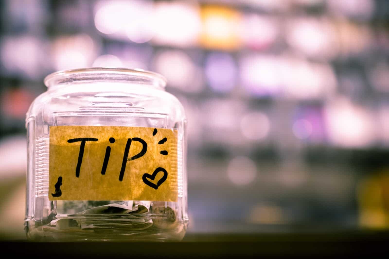 A clear glass jar with a yellow label that reads "TIP" in bold black letters, accented with small drawings of a heart and an exclamation mark. The jar contains some paper money and coins. The background is blurred and filled with colorful lights and shapes.