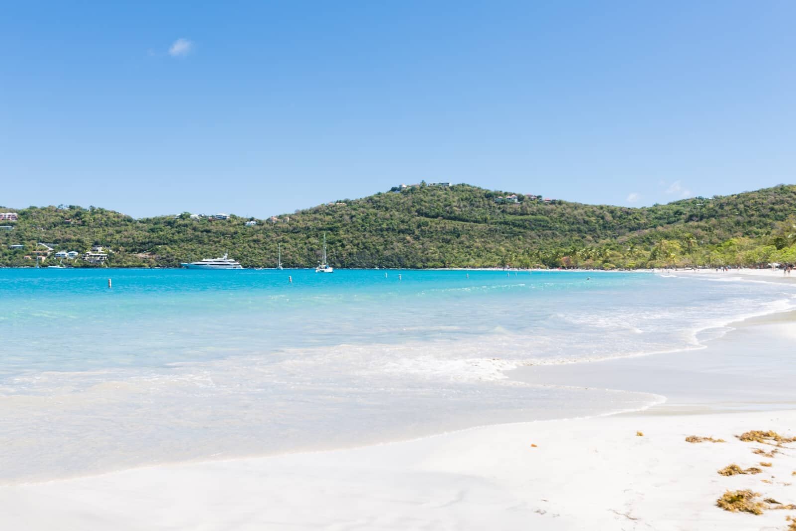 A picturesque beach with soft white sand and clear turquoise water curves along the shoreline. In the background, lush green hills rise beneath a bright blue sky. Several boats are anchored near the shore, creating a serene and inviting coastal scene.